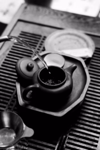 Black-and-white close-up of tea being poured from a small clay teapot on a wooden tea tray