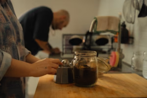 Woman's hands preparing coffee at a wooden counter, man working in the kitchen in the background