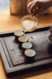Hand pouring tea from a glass pitcher into small cups arranged on a carved wooden tray