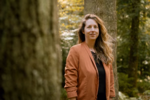 Smiling woman in an orange jacket standing among trees in a forest