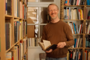 Smiling man in a brown sweater holding an open book, standing between bookshelves near a window