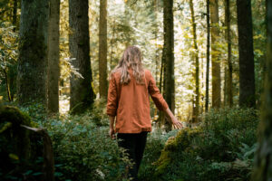 Woman in an orange top walking through forest undergrowth, seen from behind