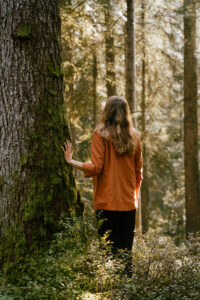 Woman in an orange top touching a moss-covered tree trunk, seen from behind in a sunlit forest