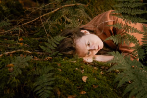 Close-up of a woman lying among ferns, eyes open, resting her head on her hand