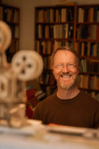 Smiling man with glasses standing in front of full bookshelves, a vintage film projector in the foreground