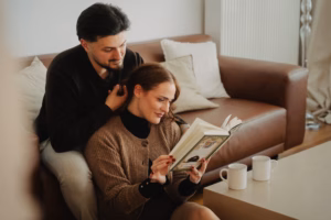 Couple sitting on a couch together, woman reading a book while the man styles her hair