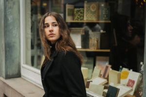 Woman with long brown hair in a black coat standing outside a bookshop window