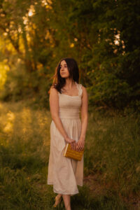 Woman in a golden-hour forest, holding a yellow book, and wearing a flowing beige dress, warm backlight through the trees.