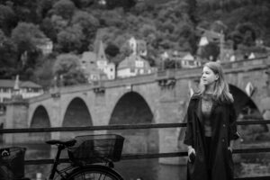 Black-and-white portrait of a woman standing by a bicycle, historic Heidelberg stone bridge in the background