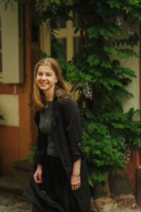 Smiling woman in a black jacket walking past a building covered in wisteria vines