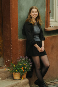 Smiling woman in a black jacket and skirt leaning against a wall next to potted flowers