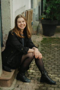 Smiling woman with long brown hair sitting on a stone doorstep, wearing a black jacket and lace-up boots, in a cobblestone alley