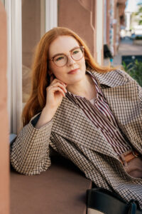 Woman with red hair and glasses in a checked coat, leaning against a window ledge with her hand near her chin