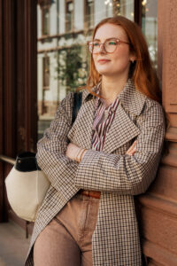 Woman with red hair and glasses leaning against a building, arms crossed, holding a cream shoulder bag