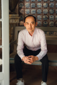 Man in a light shirt sitting on a bench, hands clasped, in a room with patterned tiled walls