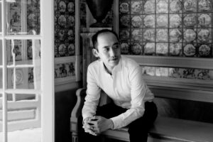 Black-and-white portrait of a man in a white shirt, sitting on a bench with hands clasped, in an ornate room with patterned tiled walls