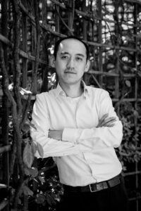 Black-and-white portrait of a man with arms crossed, standing in front of a wooden trellis