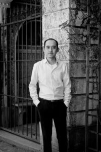 Black-and-white portrait of a man with hands in his pockets, standing beside a stone pillar and iron gate