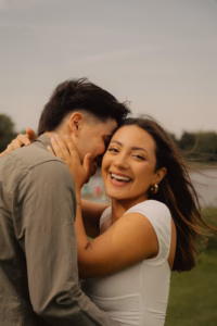 Laughing woman with her arms around her partner's neck, standing outdoors by a lake