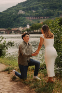 Man kneeling to propose to a woman by the Neckar riverside, town and the Heidelberg hills in the background