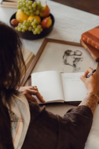 Overhead shot of a woman writing in a journal beside a bowl of fruit and a framed sketch