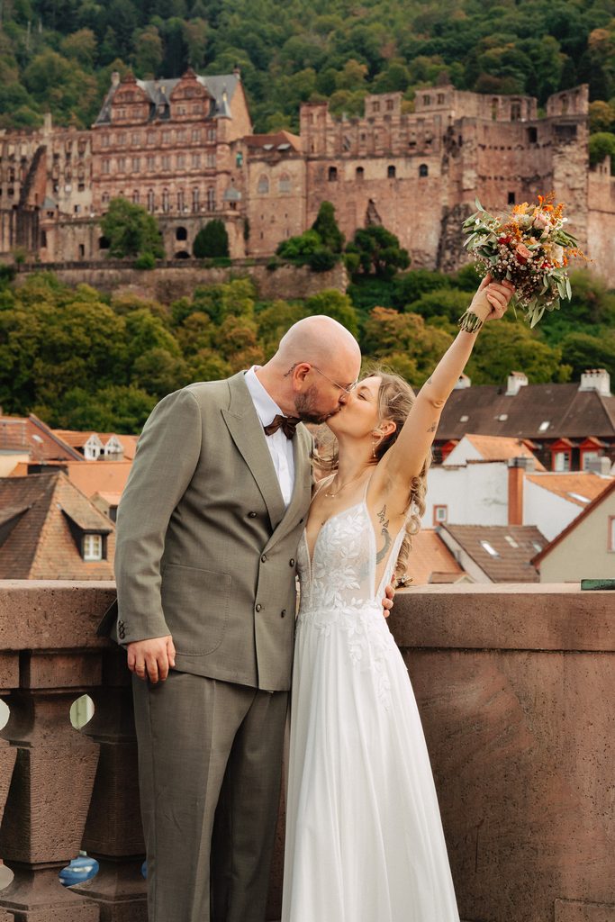 Bride and groom kissing on the Alte Brücke in Heidelberg, the bride raising her bouquet, Heidelberg Castle on the hillside behind them